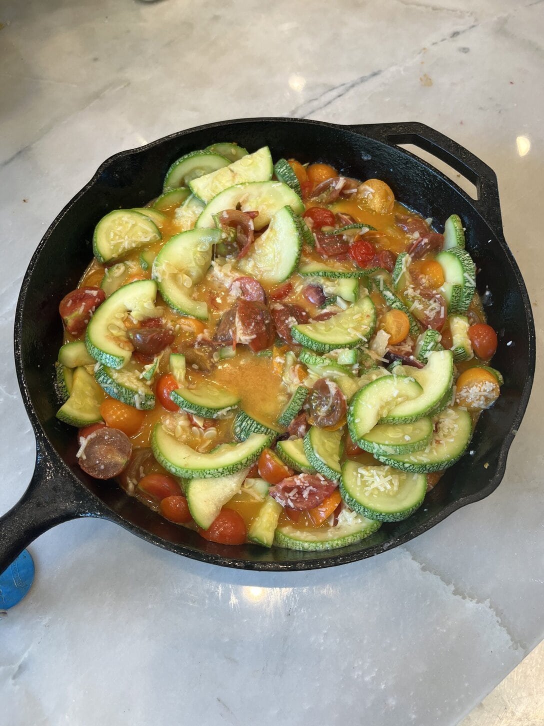 A Black Cast Iron Skillet Filled With A Colorful Vegetable Stir-Fry Featuring Sliced Zucchini, Cherry Tomatoes, And Melted Cheese, Sitting On A Marble Countertop.