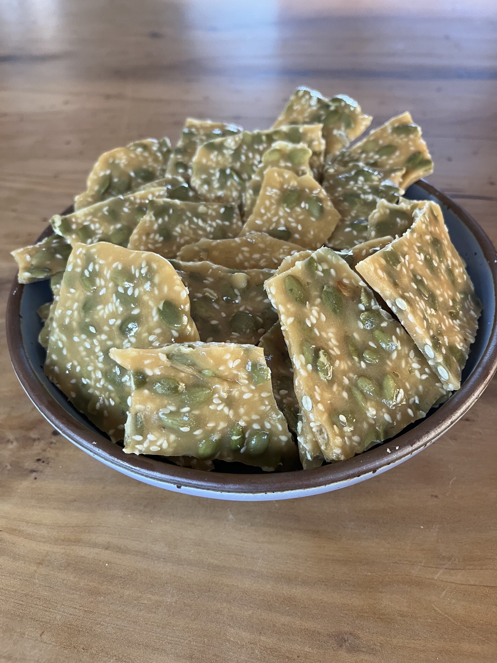 A Bowl Filled With Pieces Of Brittle Candy Containing Sesame And Pumpkin Seeds, Placed On A Wooden Surface.