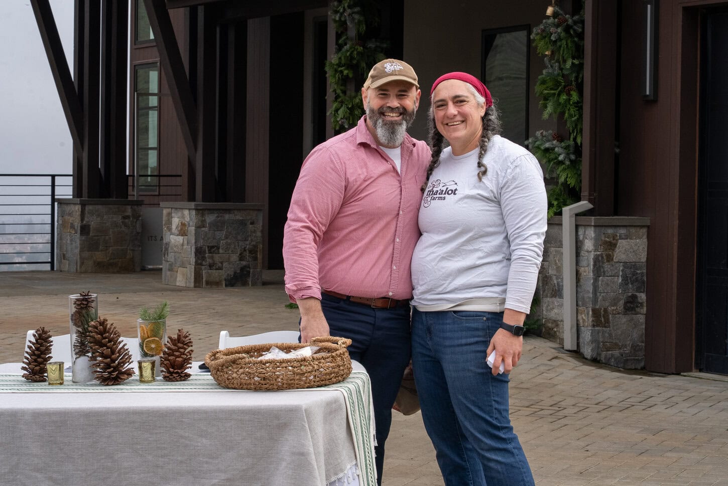 A smiling man and woman stand together outdoors beside a decorated table with pinecones and a basket. The background features a building with stone and wood details and festive greenery.