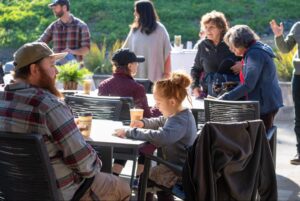 A Young Girl Sits At An Outdoor Table With Drinks, Surrounded By Adults And Other Children. Sunlight Shines On The Group As People Chat And Interact In A Relaxed Outdoor Setting.