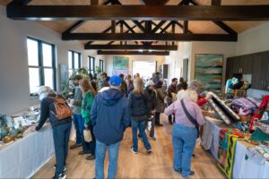 A Group Of People Browse Tables Displaying Handmade Crafts, Art, And Gifts Inside A Bright Room With Wooden Floors And Exposed Ceiling Beams During A Market Or Fair Event.
