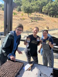 Three Women Smiling By A Table Outdoors, Holding Seed Packets And Standing Near Trays Of Soil. They Appear To Be Planting Seeds. Trees And A Dry Hillside Are Visible In The Background.
