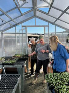 Four Adults Stand Inside A Greenhouse, Examining Trays Of Young Plants. One Person In A Gray Shirt Gestures While Talking, As The Others Attentively Observe The Seedlings Growing On Metal Tables. Sunlight Streams Through Clear Panels.
